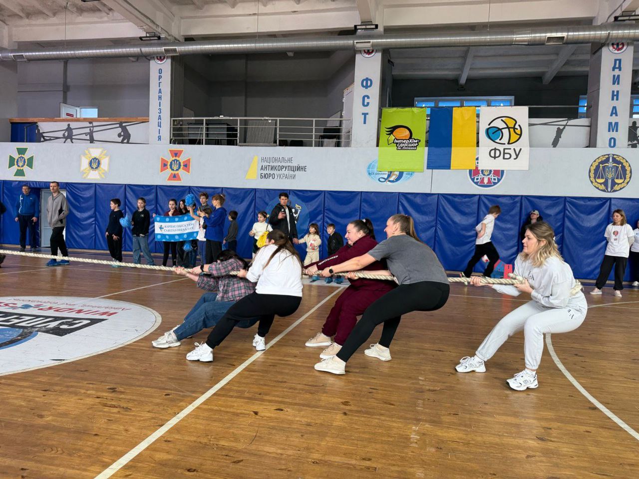 Команда жінок активно перетягує канат під час спортивних змагань у залі. На задньому плані видно вболівальників, м’які захисні мати на стінах та прапори спортивних і державних організацій.