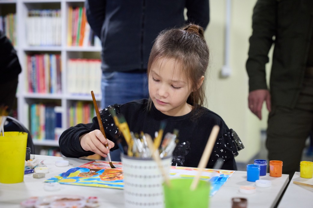 Дівчинка з темним волоссям, зібраним у хвіст, одягнена у чорний светр, зосереджено малює пензлем за столом. На столі видно пензлі у стаканчиках, фарби та її малюнок. На задньому плані розташований книжковий стелаж і стоять нечіткі силуети дорослих людей.