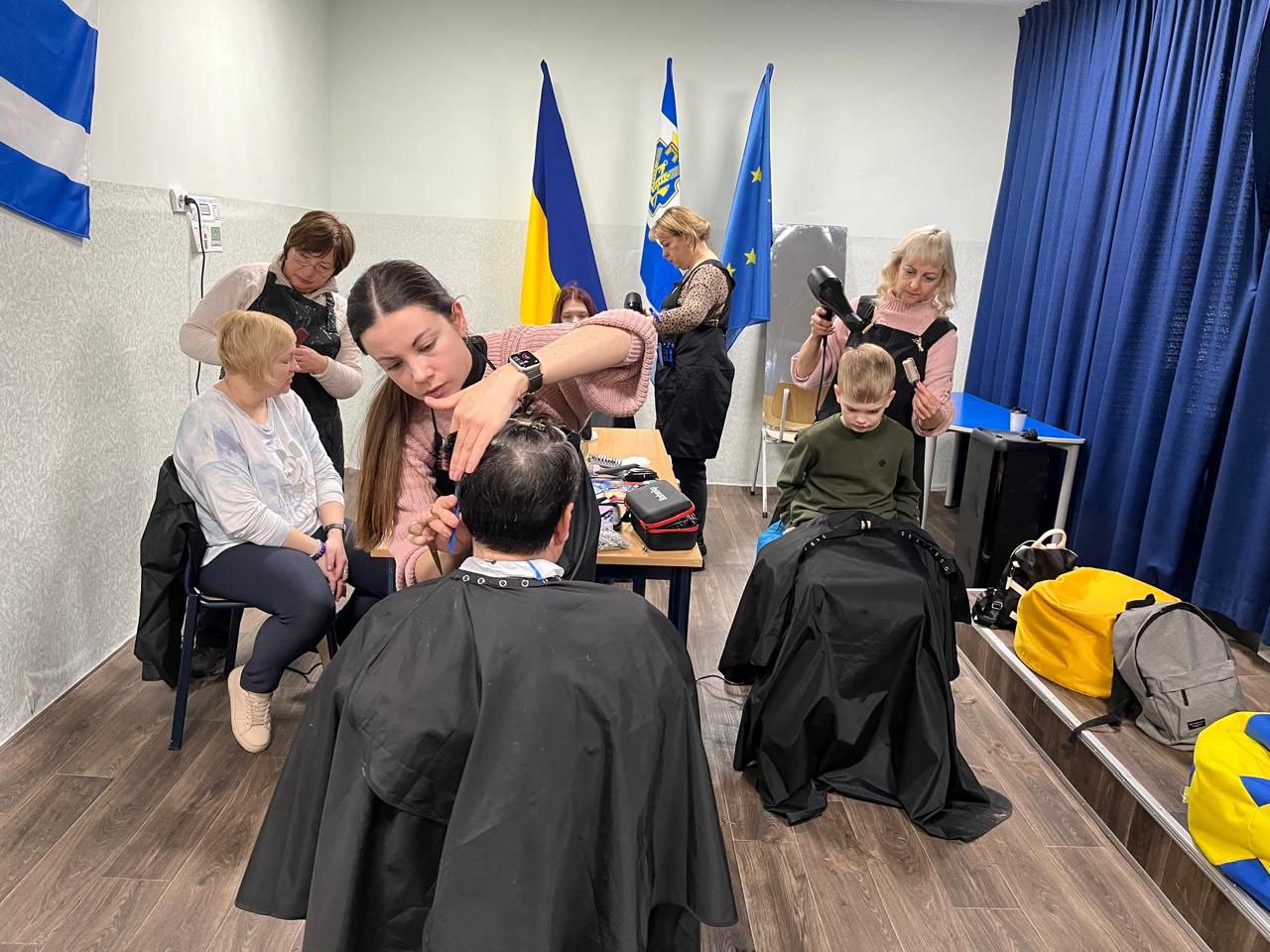 Група майстрів у приміщенні надає послуги з перукарського мистецтва дорослим та дітям. На фоні видно прапори України та Європейського Союзу.