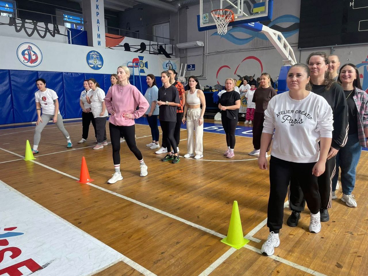 Група жінок у спортивному одязі зосереджено чекає на початок наступного етапу змагань біля розмічальних конусів на майданчику.