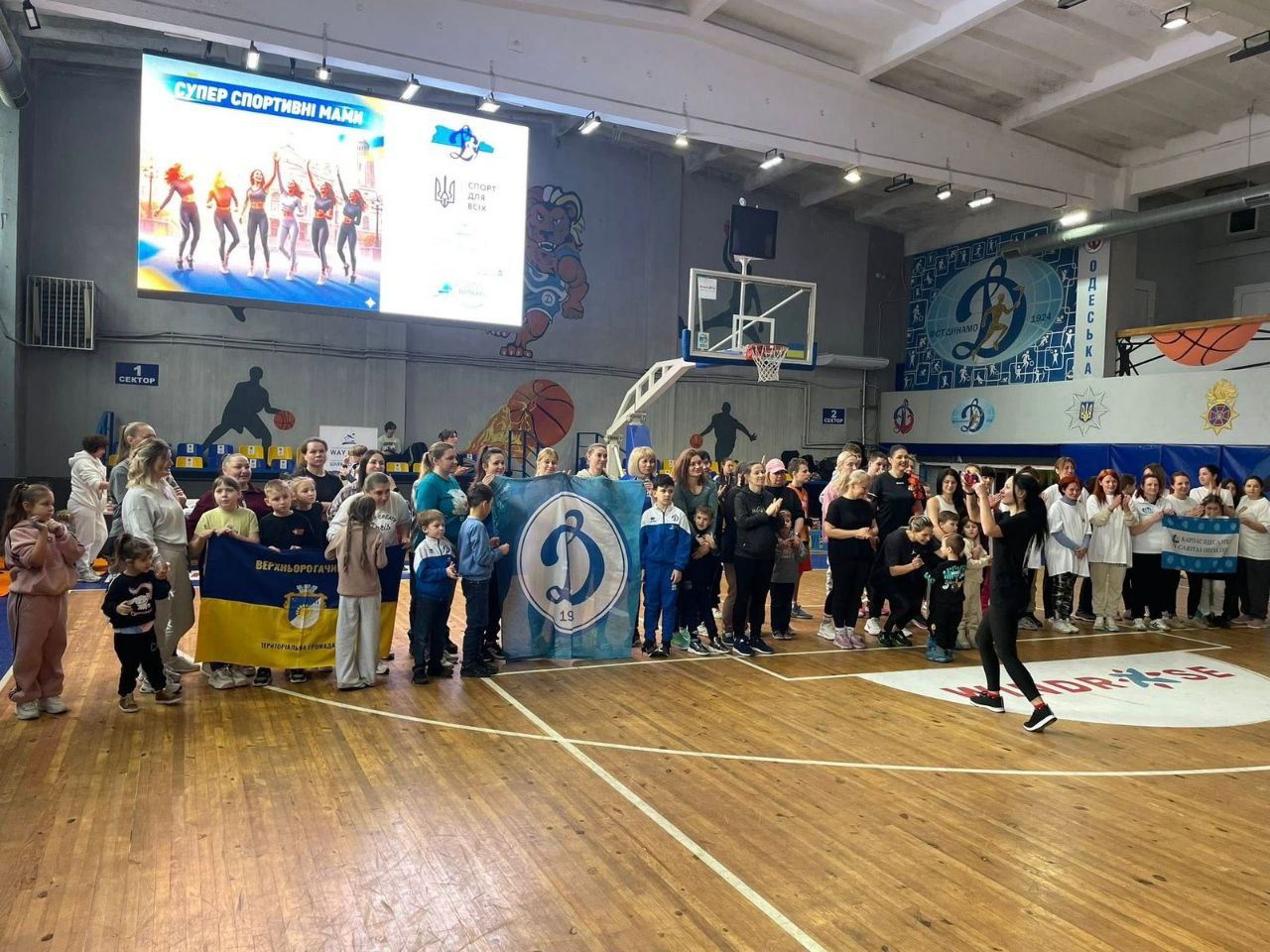 Група дорослих та дітей на спортивному майданчику під час заходу «Супер спортивні мами». Учасники тримають прапори Верхньорогачицької громади та спортивного товариства «Динамо» на фоні великого екрана та баскетбольної інфраструктури.