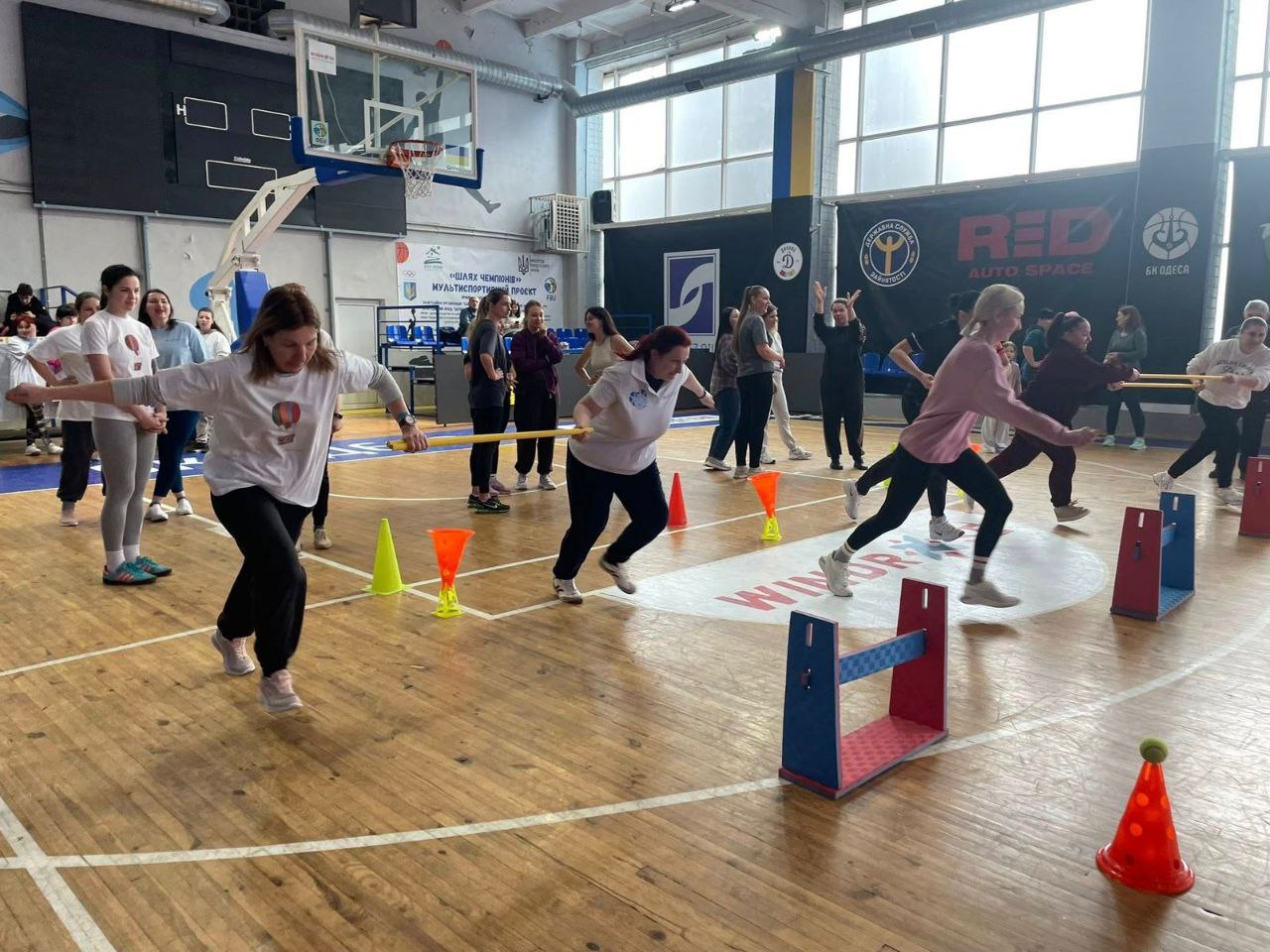 Жінки долають дистанцію з перешкодами на дерев'яному покритті залу. Учасниці перестрибують бар'єри та оббігають конуси під час спортивних змагань.
