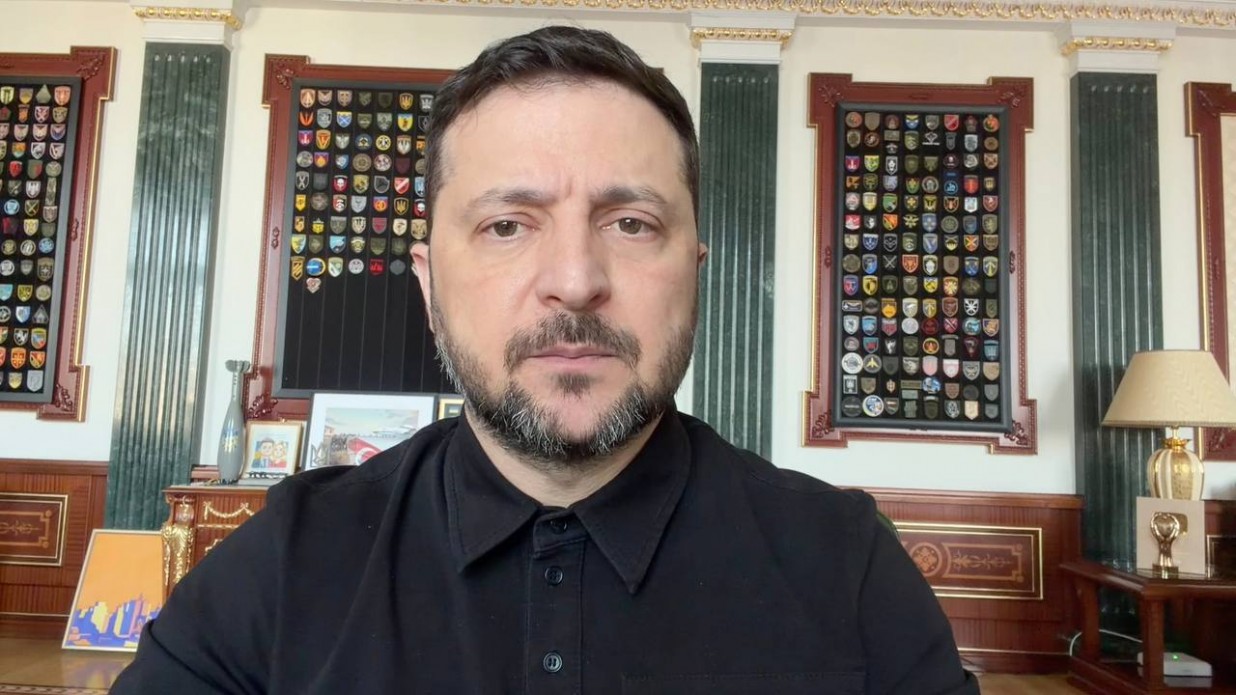 Чоловік з короткою бородою у чорній сорочці-поло звертається до аудиторії на фоні офіційного кабінету. За його спиною на стіні розміщені стенди з численними військовими шевронами та державна символіка.