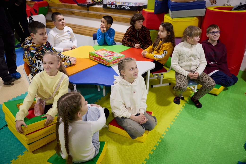 Група з десяти дітей сидить навколо яскравого різнокольорового столу в дитячій ігровій кімнаті. Деякі діти сидять на стільцях, інші – на м'яких модулях. На столі лежить коробка з грою. На задньому плані видно кольорові іграшки та модулі.