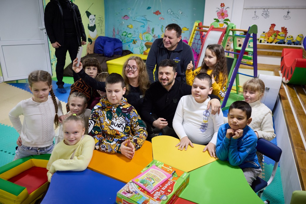Президент України Володимир Зеленський, одягнений у чорну кофту, сидить у дитячій кімнаті, оточений групою дітей і кількома дорослими. Діти та дорослі посміхаються. На столі лежить коробка з грою. На задньому плані видно яскраво розписані стіни та іграшки.