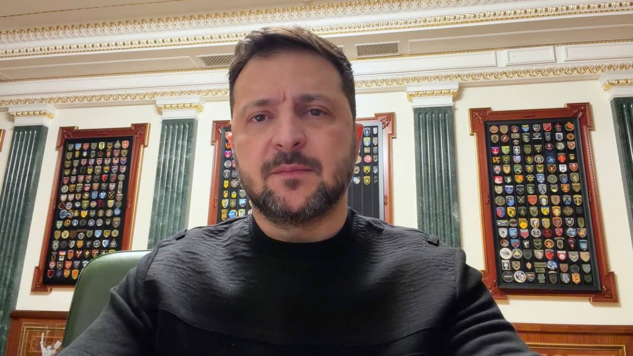 Чоловік із короткою бородою та в темному одязі на фоні офісного інтер’єру. Позаду нього на стінах розміщені декоративні стенди з численними військовими шевронами та емблемами.