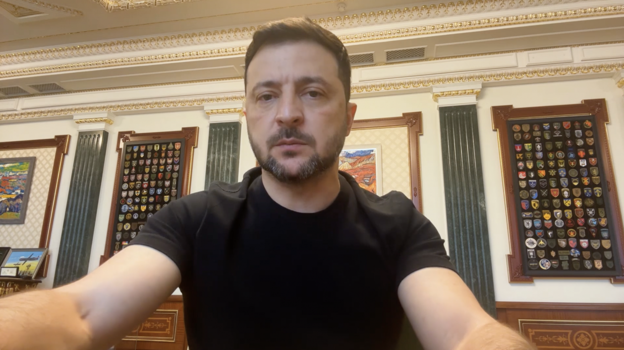 Президент України Володимир Зеленський у чорній футболці під час звернення, на тлі стіни з великими панелями, заповненими військовими нашивками та шевронами.