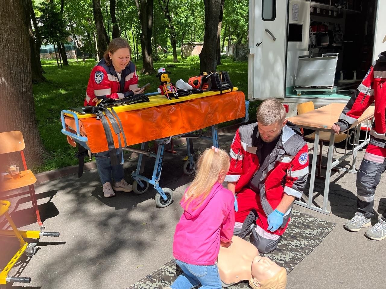 На подвір'ї біля реанімобіля медик у червоно-сірій формі екстреної медичної допомоги стоїть на колінах поруч із маленькою дівчинкою у рожевій куртці. Дівчинка відпрацьовує непрямий масаж серця на медичному манекені, що лежить на камуфляжному килимку. Позаду видно медичні ноші-каталку з помаранчевим покриттям та іншу працівницю бригади.