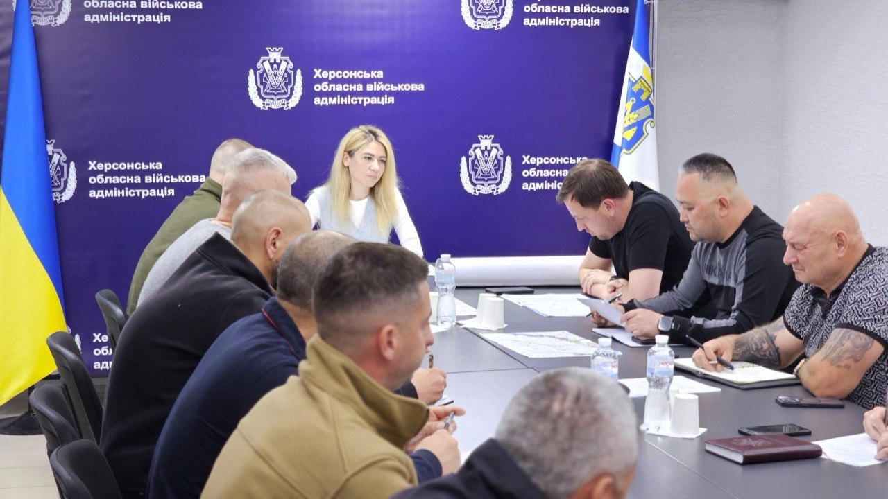Ракурс збоку на учасників наради за столом. У центрі кадру, на тлі фірмового темно-синього банера Херсонської ОВА, сидіння займає світловолоса жінка в офісному одязі, яка уважно слухає доповідачів. Праворуч від неї сидять троє чоловіків, один із яких (найближчий) має татуювання на руці та робить записи в блокноті. На передньому плані спиною до камери сидить інша частина учасників у цивільному та військовому вбранні. На столі лежать карти, документи та стоять пляшки з водою.