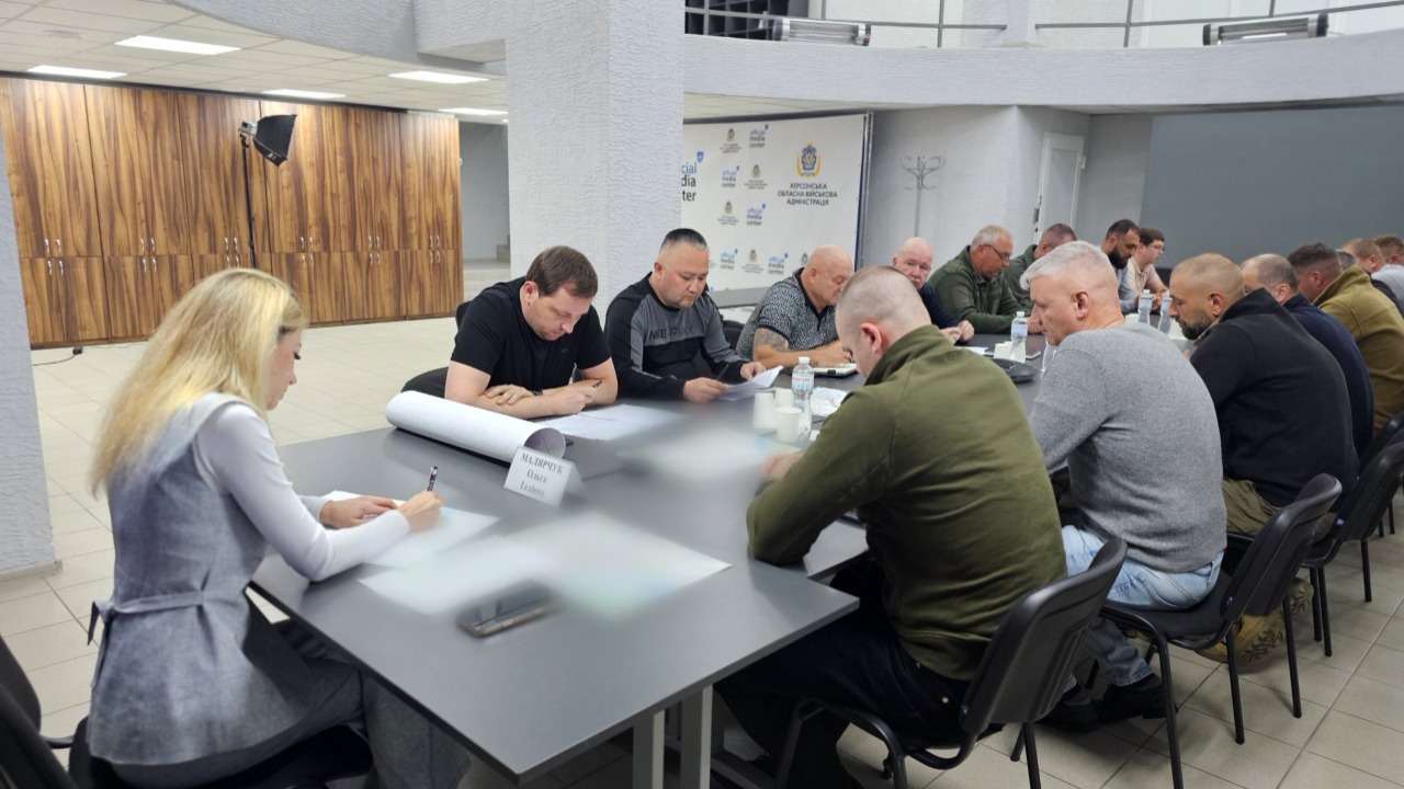 Робоча зустріч у просторій залі зі світлою підлогою. За довгим сірим столом сидить група чоловіків, які вивчають документи та роблять нотатки. Ліворуч на передньому плані сидіння займає жінка-секретар або керівник (блондинка у сірому костюмі), яка пише на аркуші паперу. Перед нею стоїть настільна табличка з написом «МАЛЯРЧУК Ольга Іллівна». На задньому плані кімнати видно дерев'яні офісні шафи, колони та мобільний стенд із логотипом ХОВА. Деякі ділянки столу та документів штучно розмиті для конфіденційності.
