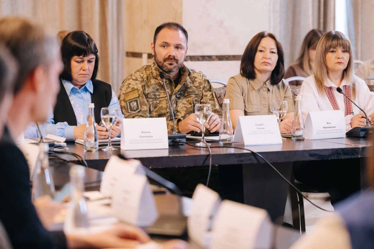 Ряд учасників сидить за довгим конференційним столом. Серед них — чоловік у військовій формі Збройних Сил України, перед яким стоїть табличка. Поруч із ним сидять жінки у повсякденному та діловому одязі, перед якими встановлено мікрофони, склянки та таблички з іменами Жанна Осипенко, Андрій Ткачов та Віта Ковальська.