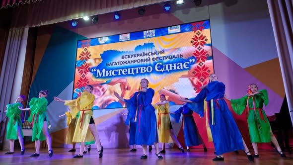 На сцені виступає група дівчат-танцівниць. Частина з них одягнена в довгі сині сукні з жовтими елементами, інші — у зелені та жовті костюми з червоними поясами. На задньому плані — великий екран із написом «Всеукраїнський багатожанровий фестиваль „Мистецтво Єднає“» на тлі карти України.