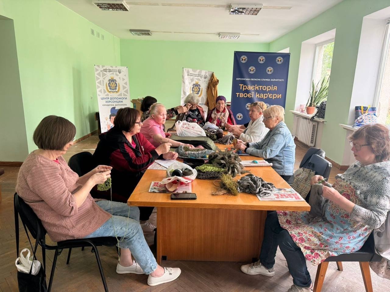 Група літніх жінок та волонтерок сидить навколо великого дерев'яного столу у світлій кімнаті з зеленими стінами. Вони займаються рукоділлям: плетуть маскувальні сітки та в'яжуть речі з текстильних стрічок і пряжі. На столі розкладені матеріали, ножиці та нитки. На задньому плані стоять білі банери «Центр допомоги херсонцям» та синій стенд Херсонської обласної служби зайнятості з написом «Траєкторія твоєї кар'єри».
