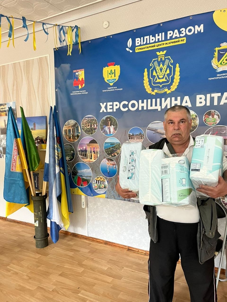 Чоловік середнього віку у білій футболці та темній жилетці тримає в руках три великі упаковки гігієнічних підгузків та пелюшок. Він стоїть у кімнаті перед великим синім стендом із круглими фотографіями краєвидів та написом «Херсонщина вітає». Ліворуч у підставці (стилізованій під гільзу) встановлені прапори України.