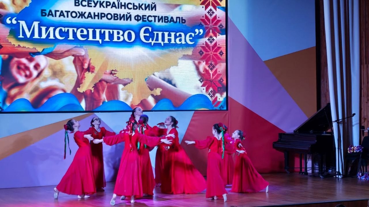 Група дівчат виконує танець, утворивши коло по центру сцени. Вони одягнені в однакові довгі червоні сукні з вплетеними у волосся стрічками. Праворуч на сцені стоїть чорний рояль. На великому екрані позаду — назва фестивалю.