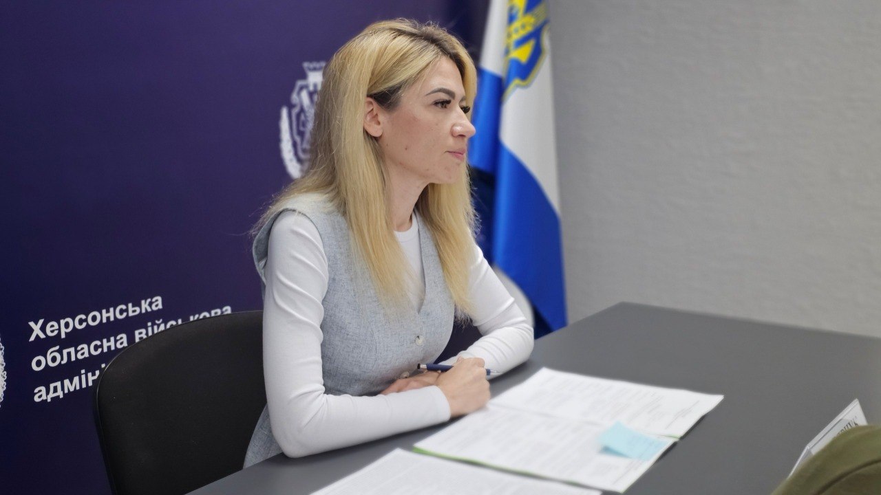 Портретний знімок великим планом. Жінка зі світлим волоссям, одягнена у білу кофту з довгим рукавом та сіру безрукавку, сидить за офісним столом у профіль. Вона тримає в руках ручку, а перед нею лежать розгорнуті папери з текстом та блакитним стікером. На задньому плані розмито видно частину синього банера з гербом та початком напису «Херсонська обласна військова...», а також державний прапор України.