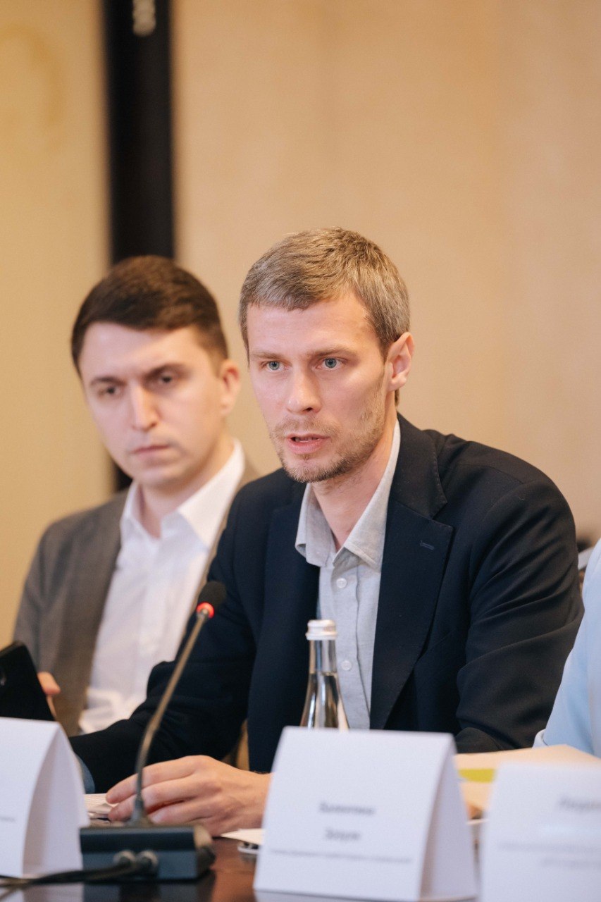 Великий план чоловіка у темному піджаку та сірій сорочці, який говорить під час обговорення. Поруч із ним, на задньому плані, сидить інший учасник заходу у сірому піджаку та білій сорочці. Перед ними на столі стоять настільні мікрофони та таблички.