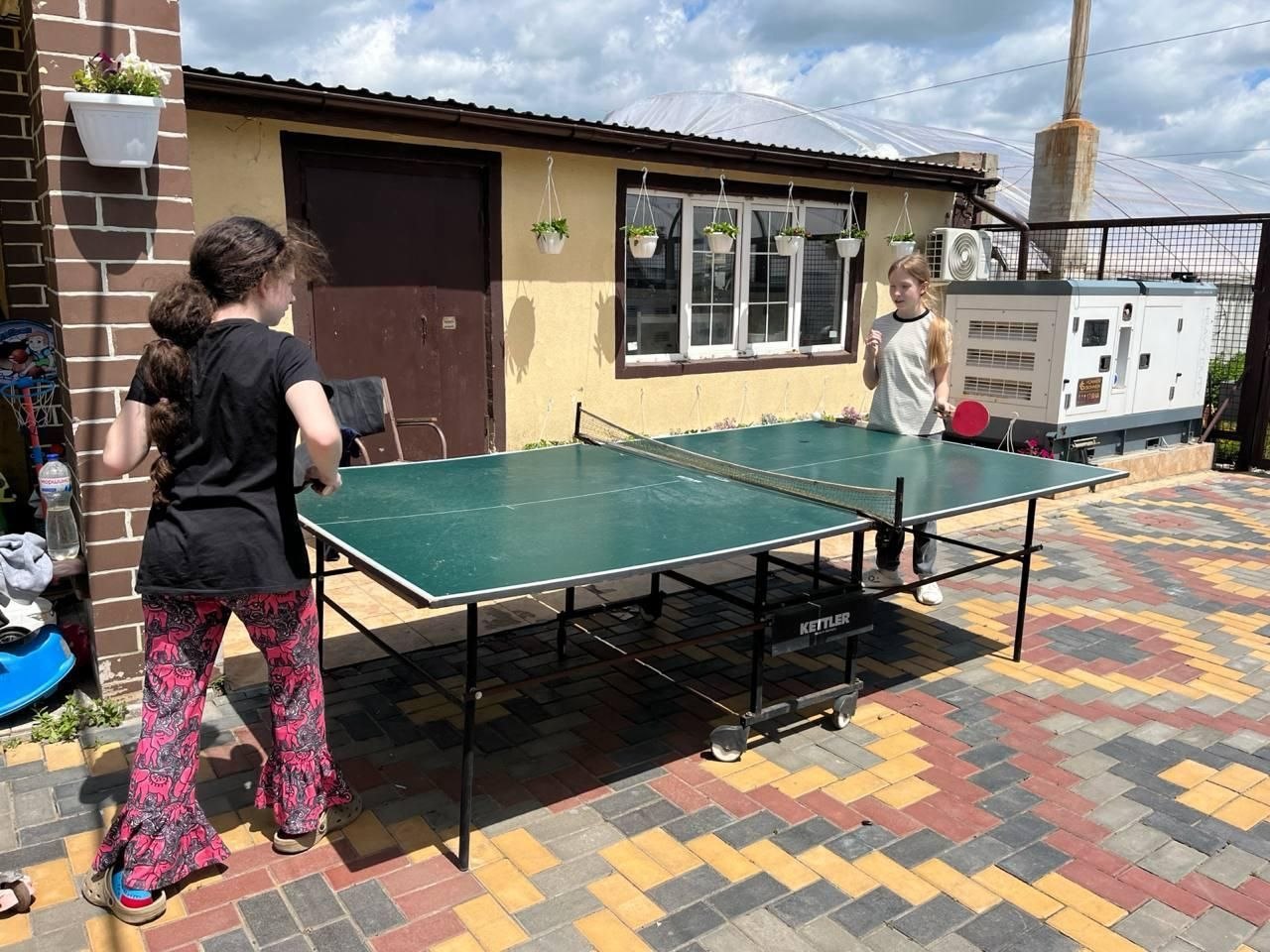 На подвір'ї, викладеному тротуарною плиткою, встановлено зелений тенісний стіл марки Kettler. Дві дівчинки грають у настільний теніс: одна стоїть спиною до кадру в яскравих рожевих штанах із візерунками, інша — з ракеткою в руці на протилежному боці столу. На задньому плані розташована одноповерхова будівля та промисловий генератор.