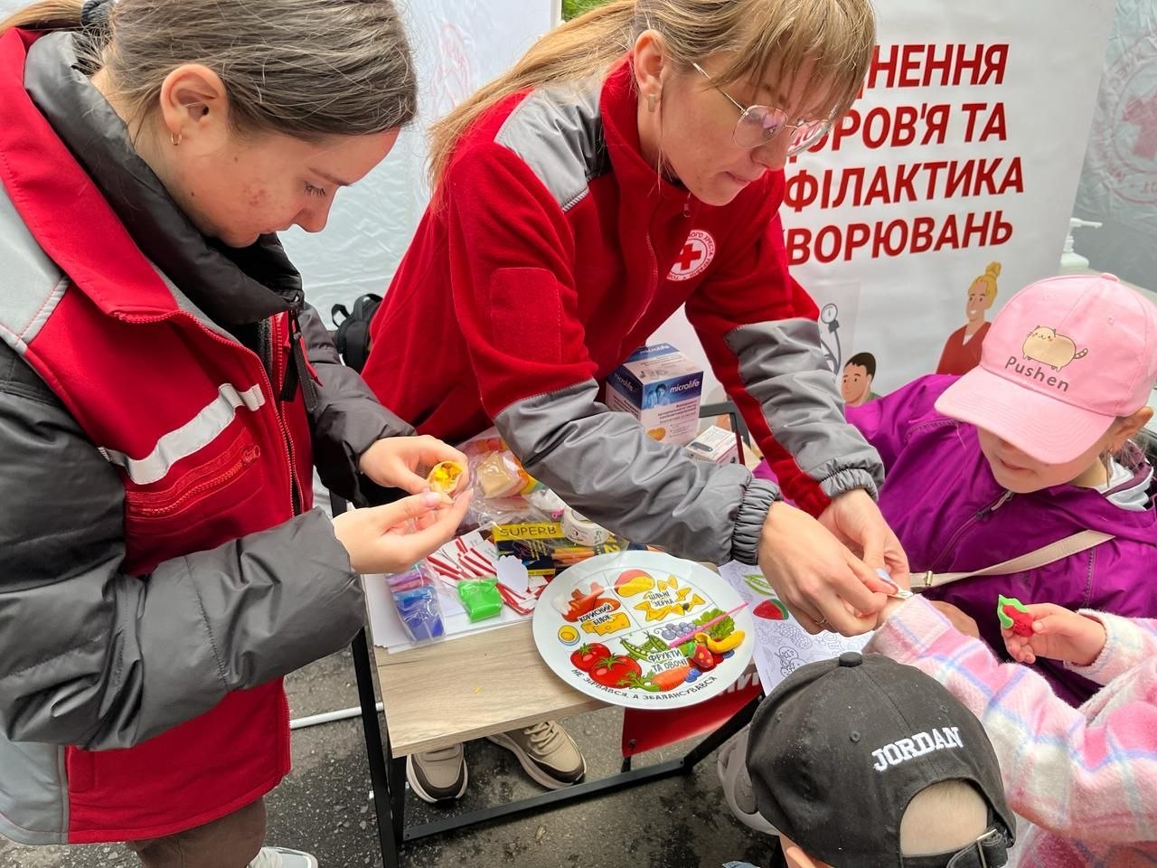 Волонтерка у червоній куртці Червоного Хреста та окулярах працює з дітьми за невеликим столом. На столі стоїть тарілка з наочним малюнком-схемою здорового раціону (овочі, фрукти, білки, злаки) та підписом «Збалансуйся». Поруч лежать матеріали для ліплення та коробка тонометра Microlife. Справа дитина у рожевій кепці «Pusheen» розглядає саморобну фігурку.