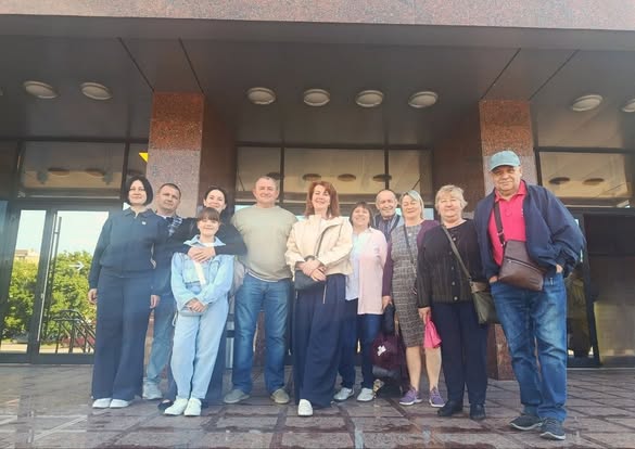 На передньому плані, на гранітному ґанку під масивним козирком будівлі, стоять одинадцять людей — чоловіки, жінки та одна дівчинка-підліток. Вони одягнені в повсякденний літній та демісезонний одяг (джинси, кофти, сорочки). Чоловік праворуч — у блакитній кепці, червоній футболці-поло та синій куртці, з коричневою сумкою через плече. Позаду них видно скляні вхідні двері.