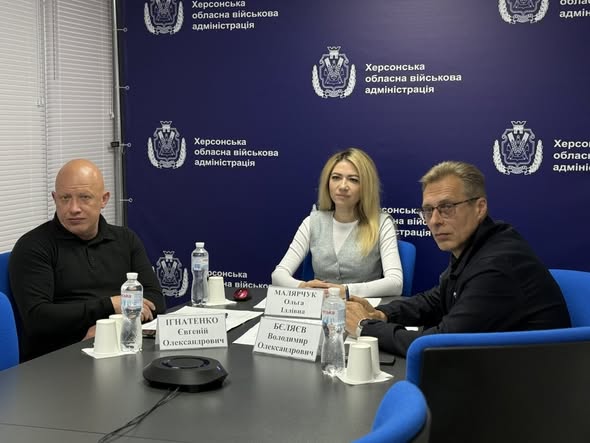 За столом у конференц-залі сидять троє людей. Ліворуч — заступник голови Херсонської ОВА Євгеній Ігнатенко у чорній сорочці-поло. По центру — заступниця голови Ольга Малярчук у світлій блузі та сірому жилеті. Праворуч — Володимир Бєляєв в окулярах та темному піджаку. Перед кожним із них на столі встановлено таблички з іменами, стоять пляшки з водою та паперові стаканчики. На задньому плані — синій брендований банер із повторюваним гербом та написом «Херсонська обласна військова адміністрація».