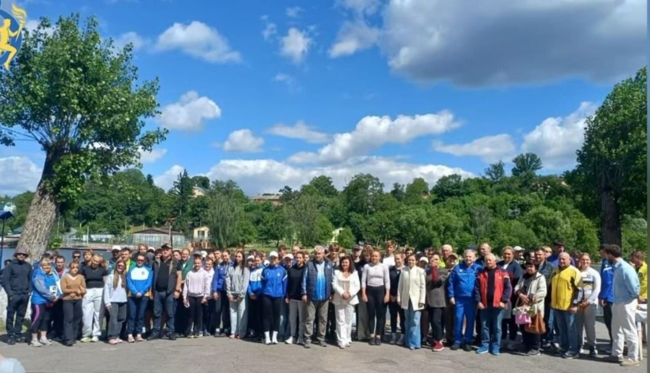 Загальний план великої групи людей (спортсмени, тренери та організатори), які вишикувалися для спільного фото на асфальтованій набережній біля води. Учасники одягнені у різноманітний спортивний та повсякденний одяг. Позаду них розгортається панорама річки та густо залісеного протилежного берега під яскраво-синім небом із пишними білими хмарами. У лівому верхньому кутку фотографії присутній жовто-блакитний логотип спортивного товариства «Динамо».