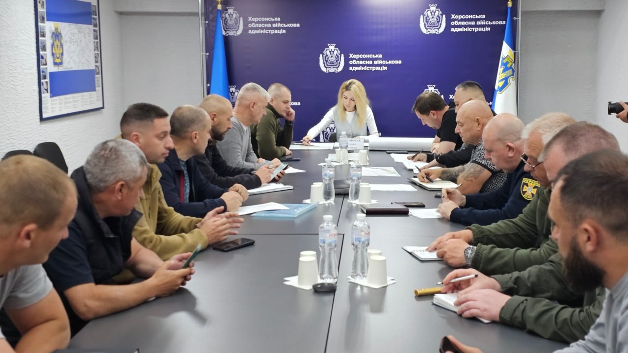 Офіційна нарада у приміщенні Херсонської обласної військової адміністрації. Великий Т-подібний стіл оточують понад десять чоловіків у військовому, спортивному та повсякденному одязі. На чолі столу сидить заступниця начальника Херсонської ОВА Ольга Малярчук у світлій жилетці поверх білої кофти, яка робить записи у документах. Перед нею встановлено табличку. Уздовж столу розставлені пляшки з водою, пластикові стаканчики та блокноти. Позаду очільниці — синій банер із символікою та повторюваним написом «Херсонська обласна військова адміністрація», по боках якого стоять державні прапори України.