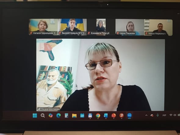 На екрані відкрито вікно відеозв'язку. У головному вікні виступає жінка в окулярах та чорній кофті, підписана як «Оксана Шестакова». Позаду неї видно малюнок із портретом Ернеста Гемінгвея на тлі книжкової полиці. У верхньому рядку відображаються інші підключені учасники: Наталія Чернишева, Валерій Крівуля (на тлі прапора України), Єлизавета Пільгуй, Ілона Пашкова та Миша Марканыч.