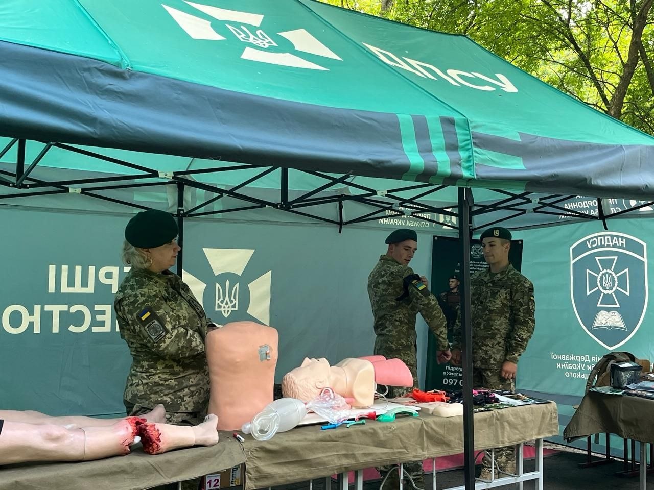 Під зеленим брендованим тентом з логотипом Державної прикордонної служби України (ДПСУ) троє військовослужбовців у камуфляжі та зелених беретах демонструють навички допомоги. На столі розкладено медичні тренажери: манекени торса людини, голови для забезпечення прохідності дихальних шляхів та модель ніг із травматичною ампутацією. Один із військових накладає турнікет на руку колезі.