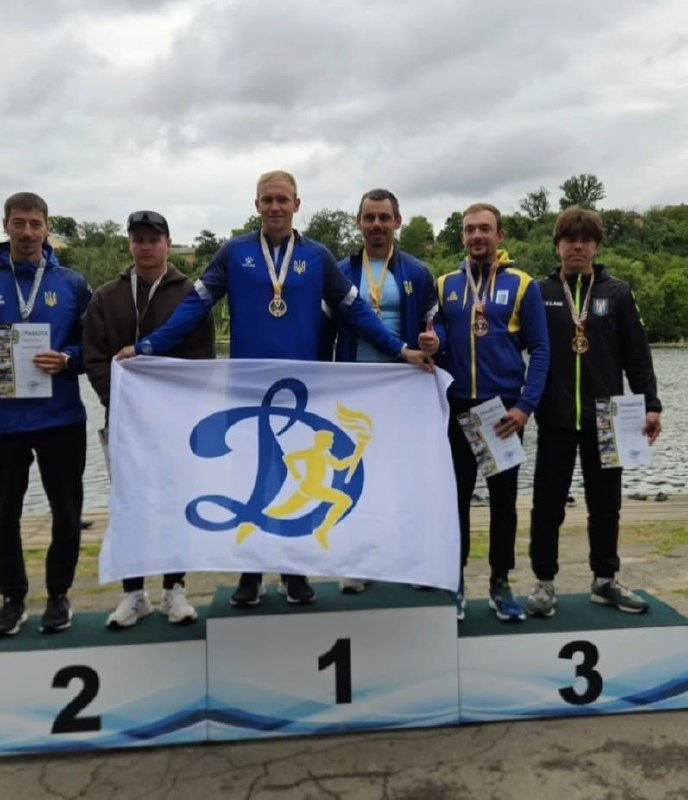 Шість спортсменів-чоловіків з медалями на шиях стоять на призовому п'єдесталі поблизу водойми. Веслувальники на першому місці тримають розгорнутий великий білий прапор із синьою літерою «Д» та емблемою бігуна з олімпійським вогнем (символіка ФСТ «Динамо»). Спортсмени на другому та третьому місцях тримають у руках залікові грамоти. На тлі видніється вода та зелений протилежний берег під хмарним небом.
