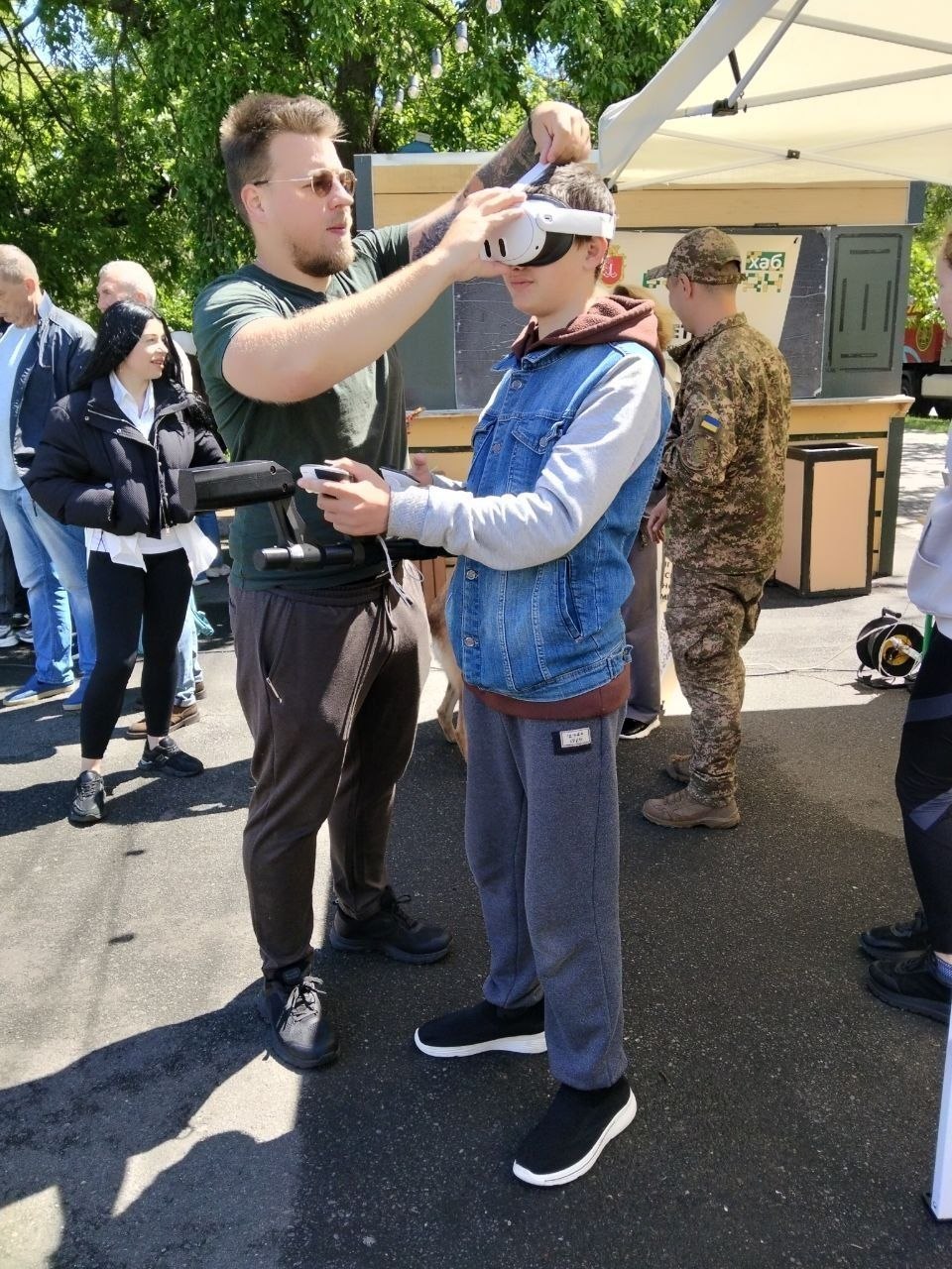 На вулиці чоловік у зеленій футболці та сонцезахисних окулярах регулює VR-гарнітуру на голові хлопця, який одягнений у джинсову жилетку та сірі спортивні штани. Підліток тримає в руках контролери. Поруч стоїть військовослужбовець у камуфляжі, а на задньому плані видно інших відвідувачів заходу.