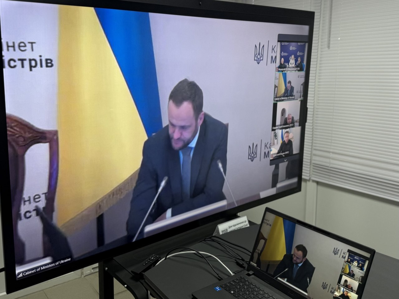Робоче місце для відеоконференцій. На великому моніторі та на екрані ноутбука, що стоїть поруч, транслюється хід онлайн-засідання. Головне вікно показує виступ посадовця у костюмі та краватці на тлі прапора України та логотипа Секретаріату Кабінету Міністрів. У правій частині екрана видно мініатюри інших підключених учасників, зокрема вікно представників Херсонської ОВА.