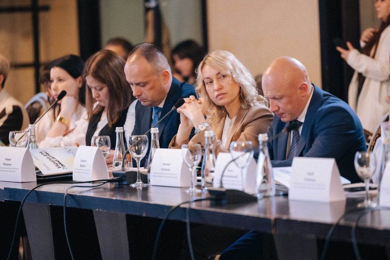 Група з п'яти учасників сидить за офіційним столом під час засідання. Серед них — чоловік у синьому піджаку, жінка з русявим волоссям у бежевому жакеті, чоловік у білій сорочці та інші представники. Перед ними розставлено настільні мікрофони, келихи та таблички, зокрема з іменами Роман Романюк та Людмила Бурдіна.