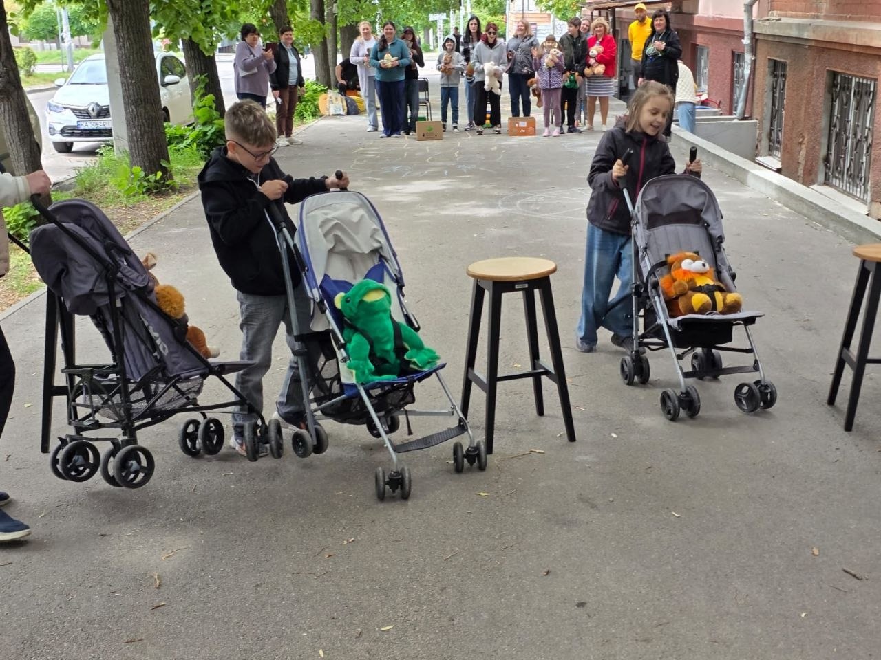 На асфальтованій доріжці біля будівлі двоє дітей — хлопчик в окулярах та дівчинка — на швидкість штовхають перед собою дитячі прогулянкові візочки сірого кольору. У візочках замість дітей пристебнуті великі м'які іграшки (зелена жаба та помаранчевий ведмідь). Між ними стоїть дерев'яний стілець-табурет, що слугує орієнтиром. На задньому плані за змаганнями спостерігає група дорослих.