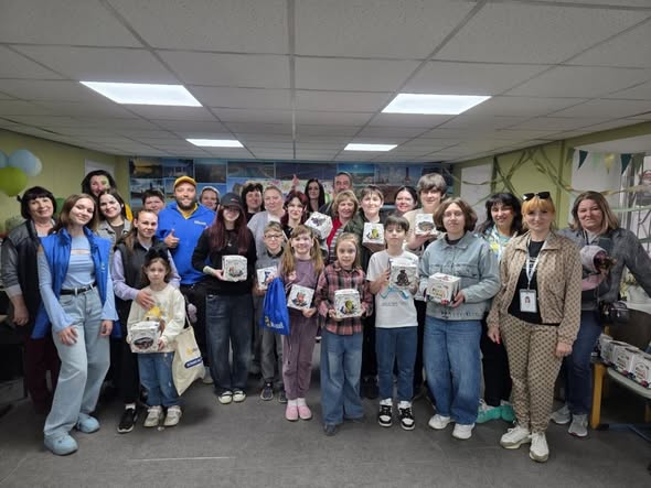 У приміщенні з підвісною стелею позує група з понад двадцяти людей різного віку — діти, підлітки та дорослі. Багато з них тримають у руках однакові невеликі квадратні коробки з малюнками. Зліва стоїть дівчина у синій жилетці поверх блакитного топа. Деякі учасники посміхаються. На задньому плані стіна прикрашена колажем із фотографій.