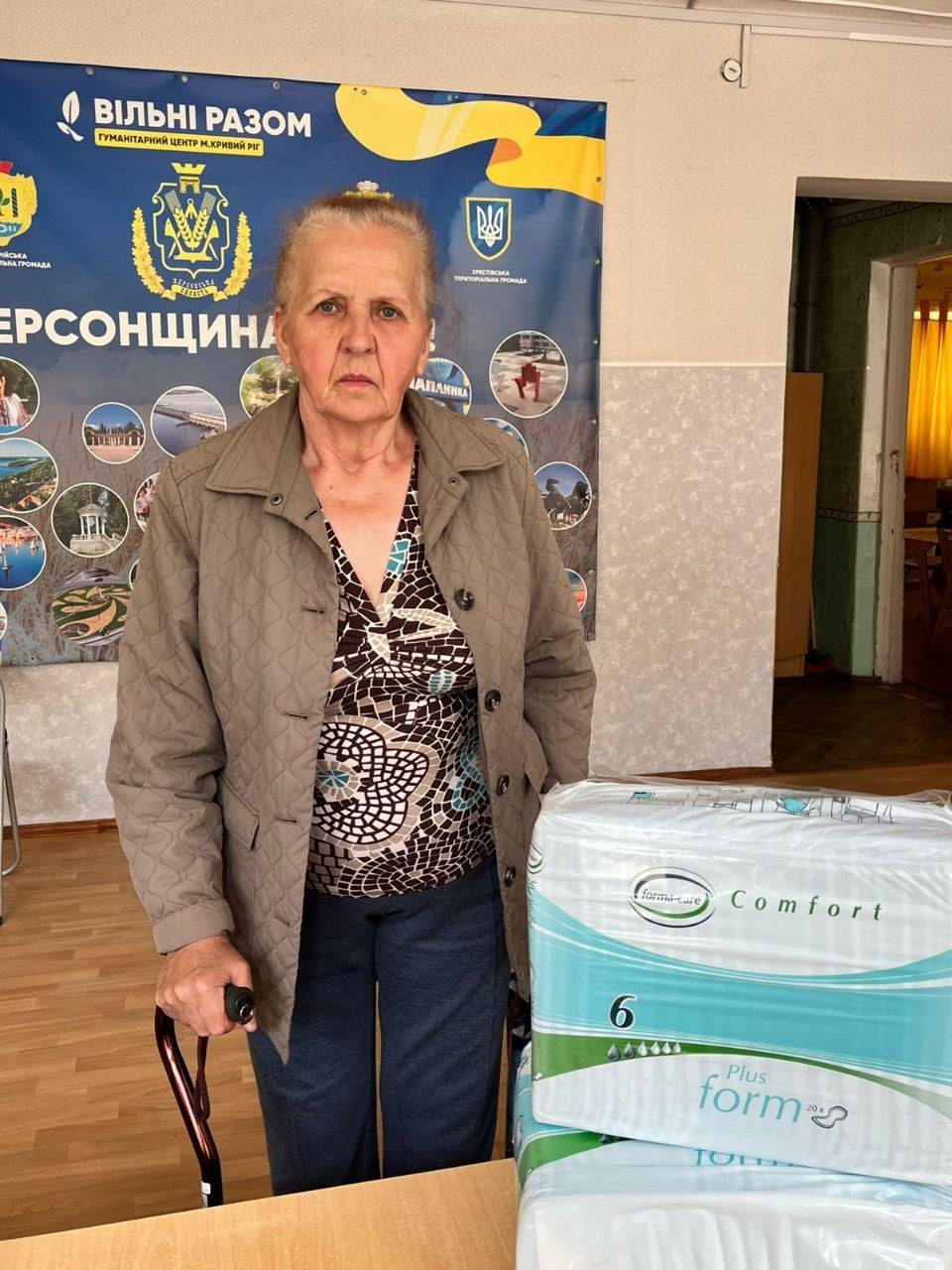 У приміщенні хабу стоїть літня жінка у коричневій стьобаній куртці, спираючись рукою на тростину. Поруч на столі лежить велика упаковка засобів гігієни бренду Forma-care Comfort (розмір 6). На задньому плані — синій банер із символікою громадської організації, гербом України та написом «Херсонщина».