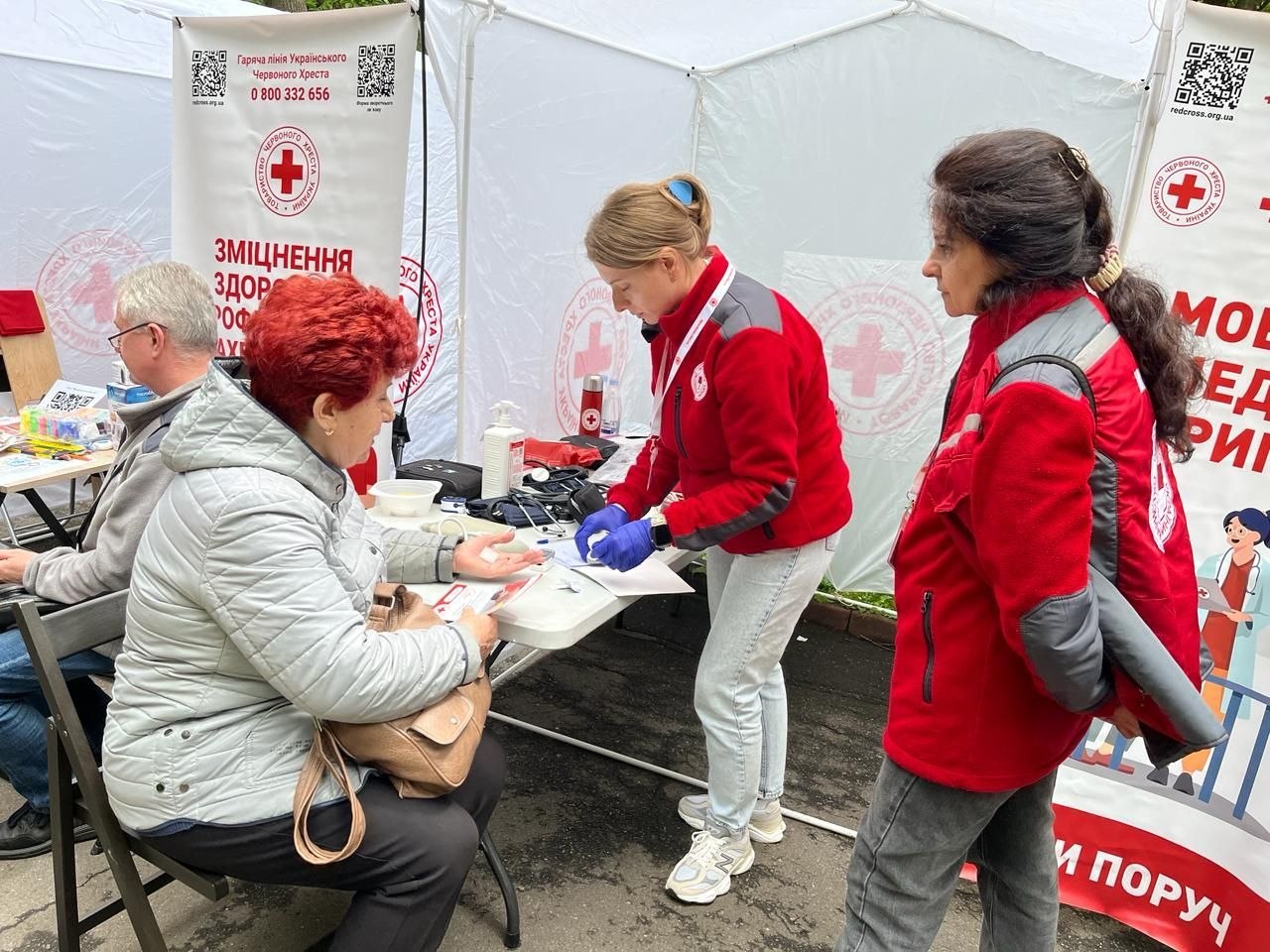 У білому наметі медична працівниця у червоній фірмовій куртці та синіх гумових рукавичках фіксує манжету тонометра на руці літньої жінки з рудим волоссям. Поруч стоїть ще одна співробітниця організації. На задньому плані видно інформаційні банери з емблемою Червоного Хреста, номером гарячої лінії 0 800 332 656 та написом «Мобільні медичні бригади».