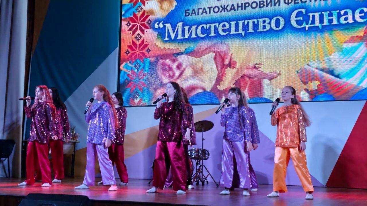 П'ять дівчат-вокалісток співають у мікрофони на сцені. Вони одягнені в яскраві блискучі костюми (паєткові кофти та вільні штани) бордового, фіолетового, бузкового та помаранчевого кольорів. На задньому плані видно частину ударної установки та великий екран фестивалю.