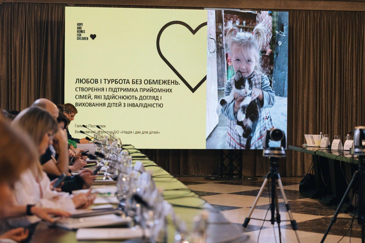 На великому екрані у конференц-залі відображається презентаційний слайд світло-жовтого кольору. У лівій частині розміщено логотип організації «Hope and Homes for Children», графічне зображення серця та чорний текст: «ЛЮБОВ І ТУРБОТА БЕЗ ОБМЕЖЕНЬ. СТВОРЕННЯ І ПІДТРИМКА ПРИЙОМНИХ СІМЕЙ, ЯКІ ЗДІЙСНЮЮТЬ ДОГЛЯД І ВИХОВАННЯ ДІТЕЙ З ІНВАЛІДНІСТЮ». Нижче вказано ім'я доповідача: Галина Постолюк. У правій частині слайда розміщено вертикальне чорно-біле фото маленької дівчинки з двома хвостиками, яка тримає на руках кошеня. На передньому плані видно довгий стіл із мікрофонами та обладнанням у перспективі.