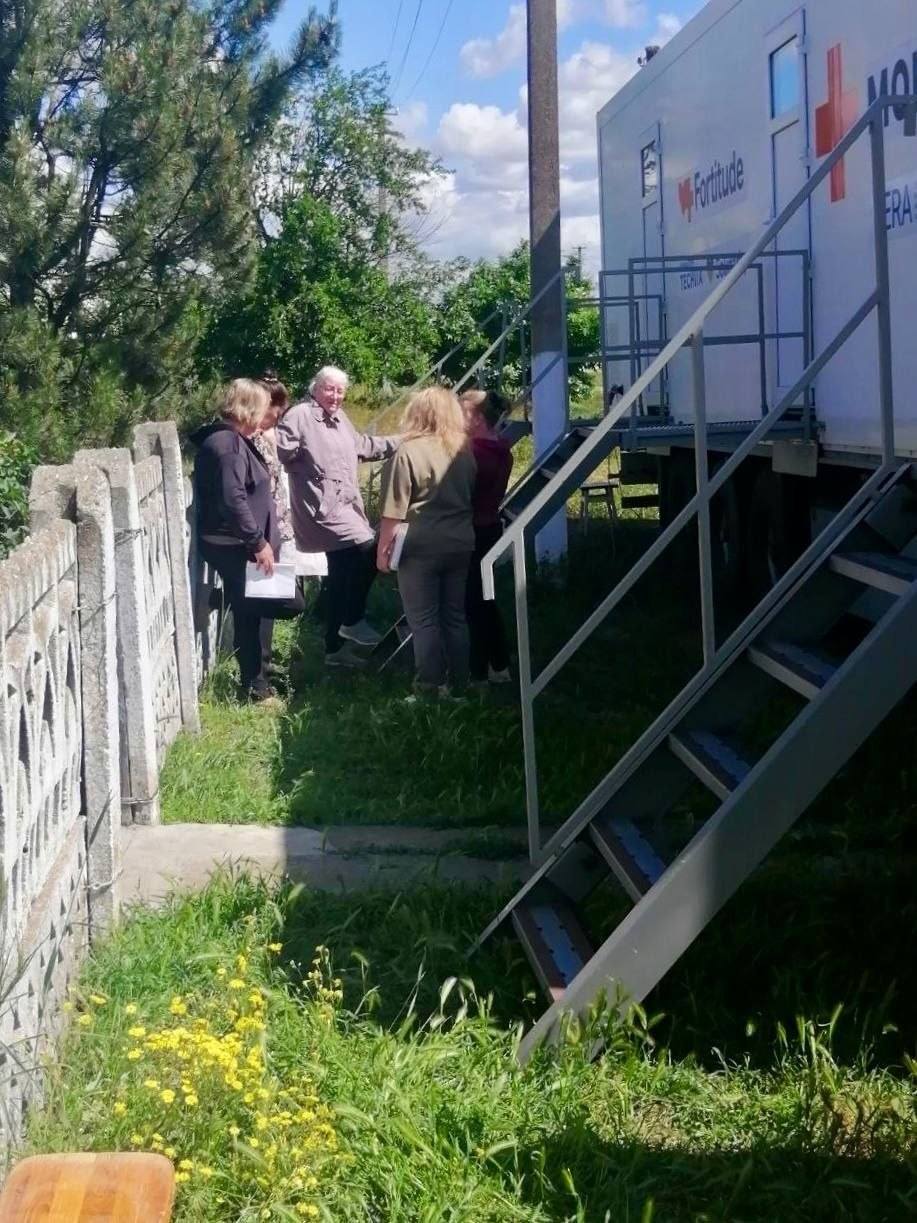 Група з чотирьох жінок спілкується на подвір'ї біля металевих сходів, що ведуть у мобільний медичний комплекс. Одна з жінок тримає в руках документи. На білому борту спецтранспорту видно логотип благодійної організації та напис «Fortitude». Ліворуч розташований бетонний паркан, на передньому плані — зелена трава з жовтими польовими квітами.