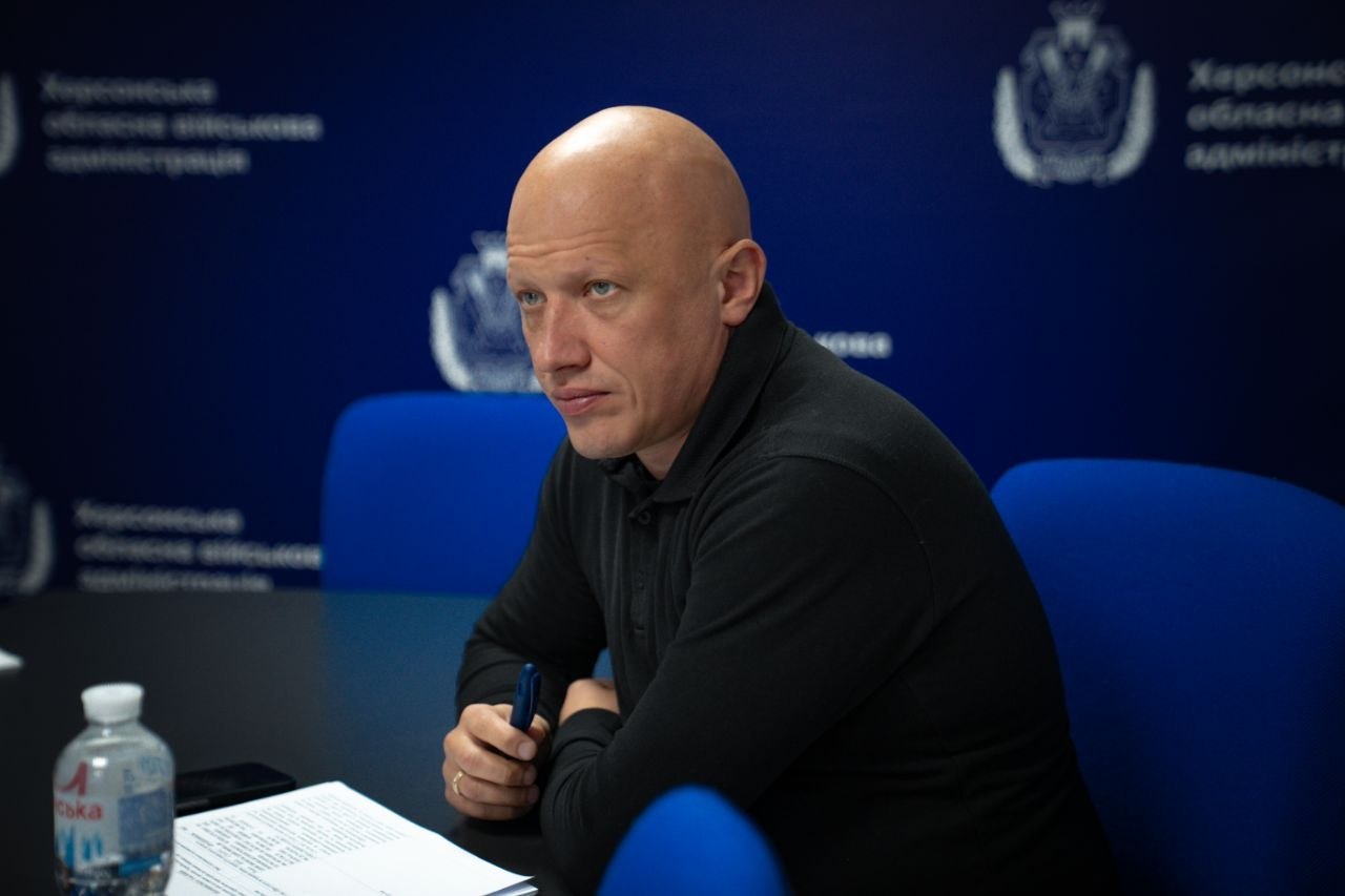 Портретний знімок збоку. Лисий чоловік (Євгеній Ігнатенко) у чорному поло з довгим рукавом сидить за столом під час робочої наради. Він зосереджено дивиться вбік, тримаючи в руці синю ручку. Перед ним на столі лежать роздруковані документи та стоїть пластикова пляшка з водою. На задньому плані — темно-синій банер із фірмовим логотипом та повторюваним написом «Херсонська обласна військова адміністрація», а також порожні сині офісні крісла.