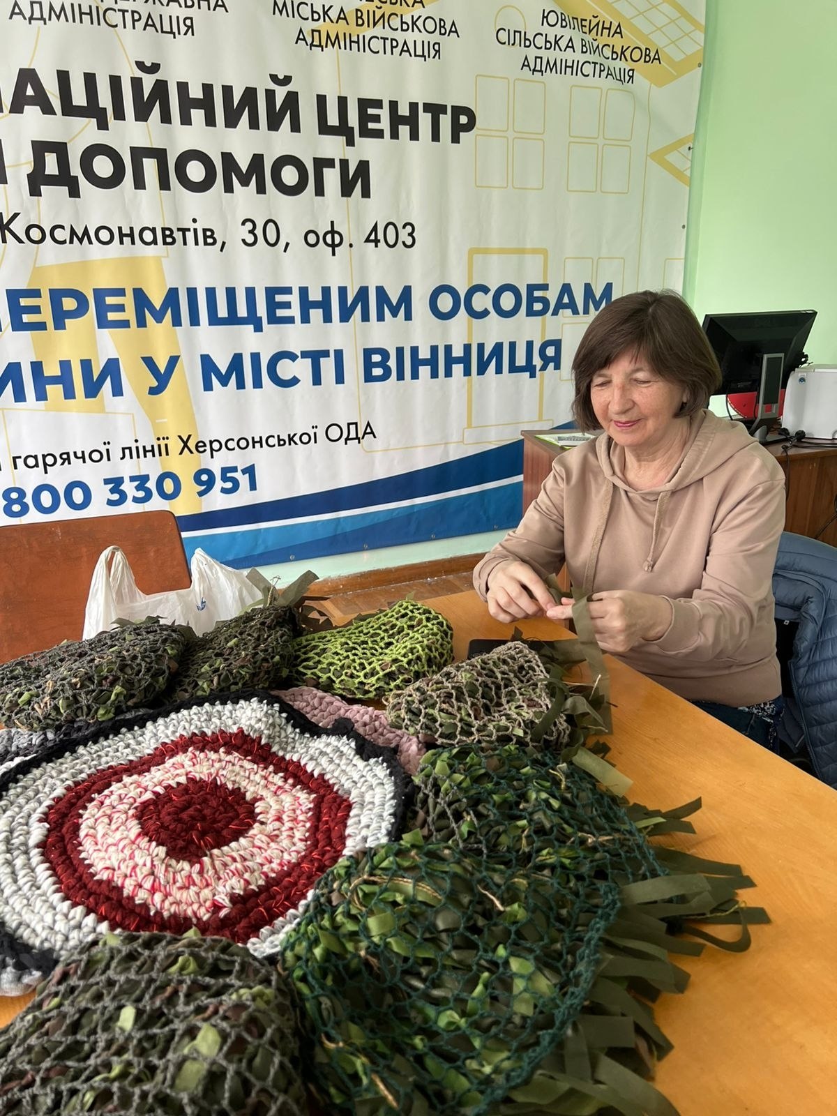 Жінка у бежевому худі сидить за столом і зосереджено плете маскувальний виріб. На столі перед нею розкладено кілька невеликих готових маскувальних сіток зеленого та захисного кольорів, а також круглий плетений килимок із білих, червоних і сірих ниток. Позаду неї розташований великий інформаційний банер гуманітарного центру для внутрішньо переміщених осіб із Херсонщини у місті Вінниця, де вказана адреса (Космонавтів, 30) та телефон гарячої лінії.