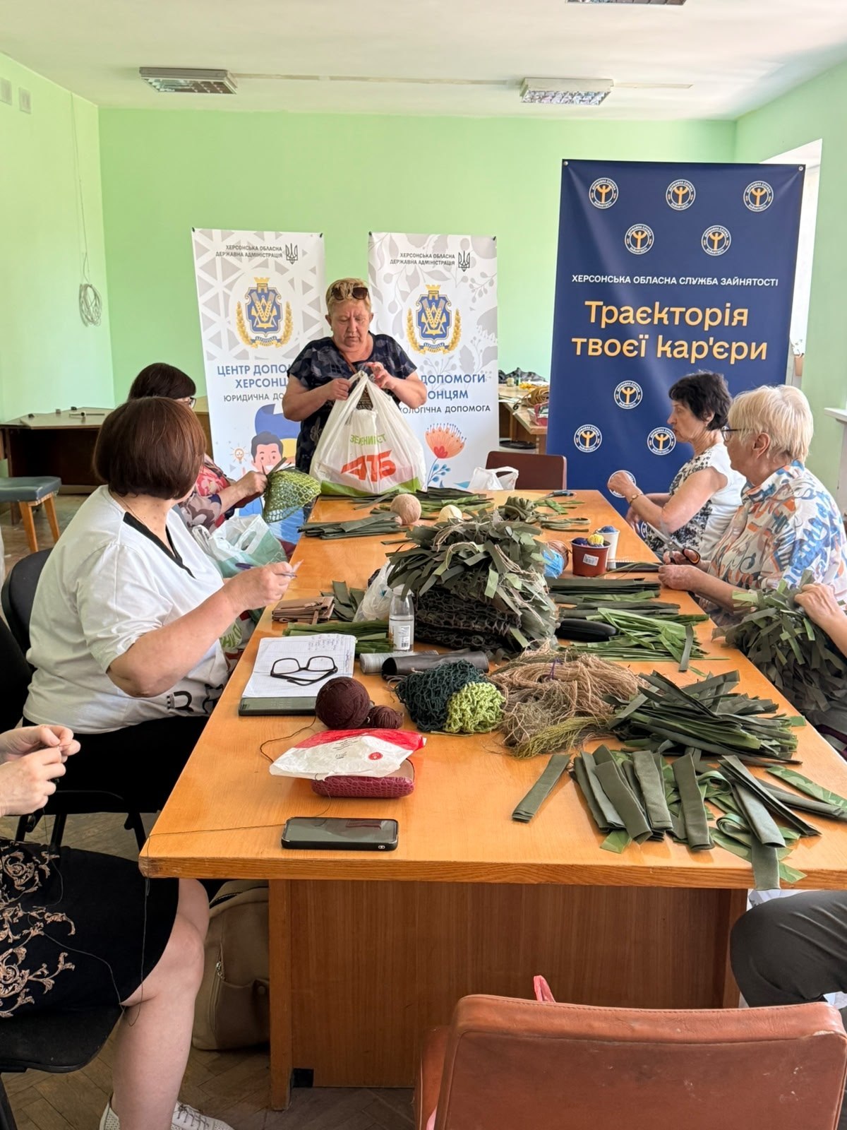 Жінки працюють за великим столом, заваленим матеріалами для створення маскувальних сіток і накидок. Одна з учасниць стоїть і дістає зелені тканинні смужки з великого пакета, інші сидять і вплітають стрічки захисних відтінків (хакі, оливковий) у сітчасту основу. На задньому плані видно брендовані банери ХОВА та служби зайнятості.