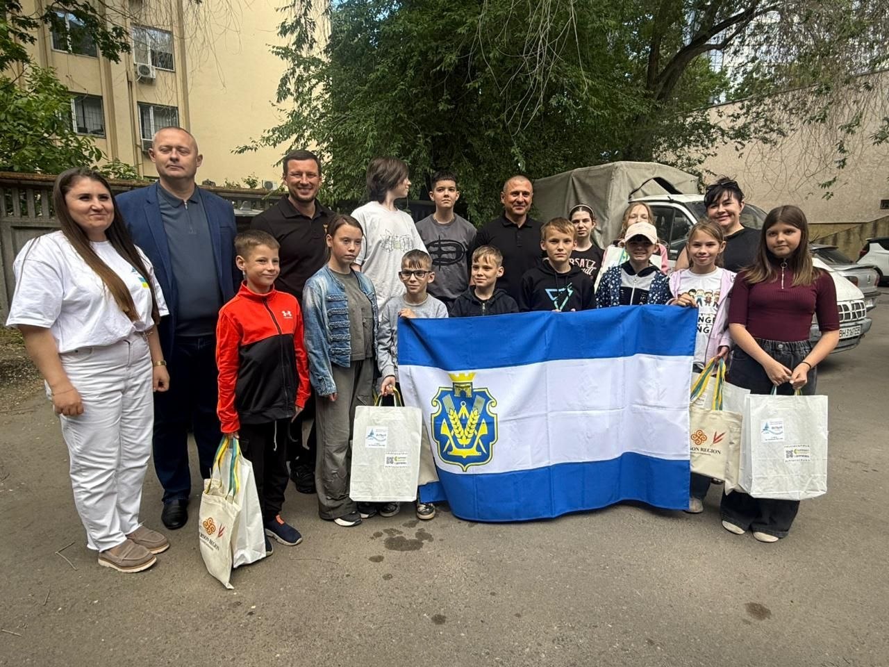 На подвір'ї будівлі стоїть група дітей різного віку та четверо дорослих. По центру діти тримають синьо-біло-синій прапор із гербом Херсонської області. Дехто з присутніх тримає екоторби з логотипами. На задньому плані видно зелені дерева та автомобіль.