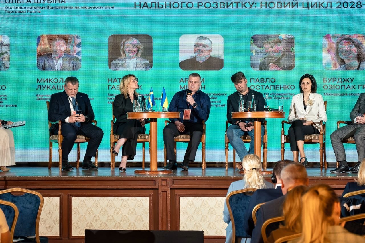 Сцена під час панельної дискусії на конференції. На дерев'яному підвищенні у кріслах сидять шестеро спікерів (чотири чоловіки та дві жінки) в офісному та повсякденному одязі. По центру чоловік говорить у мікрофон, між кріслами стоять круглі столики з пляшками води та маленькими прапорцями України і Швеції. Позаду них — величезний світлодіодний екран бірюзового кольору, на якому виведені імена доповідачів (видно написи Микола Лукашук, Павло Остапенко) та заголовок «...НАЛЬНОГО РОЗВИТКУ: НОВИЙ ЦИКЛ 2028-...». На передньому плані зі спини видно голови глядачів у залі, які слухають виступи.