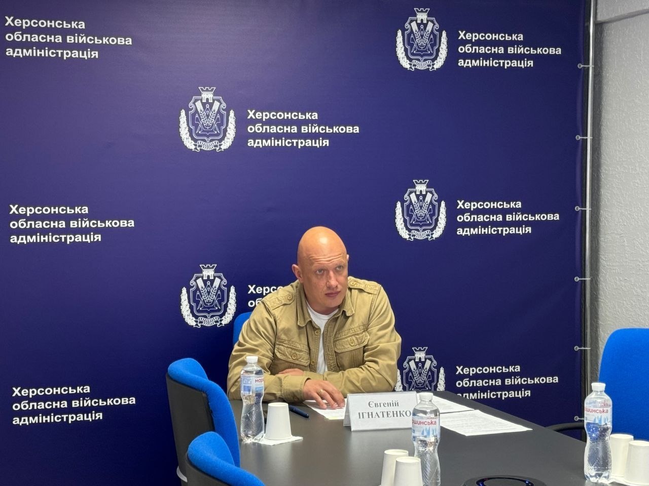Лисий чоловік у бежевій куртці-сорочці військового стилю поверх білої футболки сидить за столом під час засідання. Він дивиться вбік, спершись руками на стіл, де перед ним лежать документи, ручка та іменна табличка з написом «Євгеній ІГНАТЕНКО». На столі також стоять пластикові пляшки з водою та паперові стаканчики. Позаду нього — великий темно-синій банер (брендмауер) із повторюваним логотипом та написом «Херсонська обласна військова адміністрація». Стіл оточують сині офісні крісла.