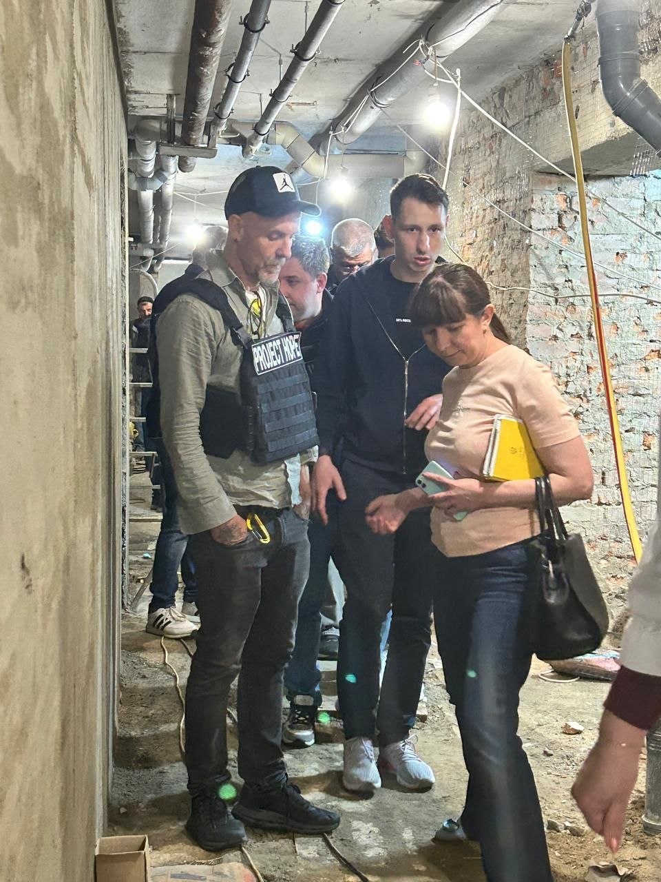 План у повний зріст. На передньому плані зліва стоїть чоловік у чорній кепці, сірій сорочці та захисному бронежилеті з чітким маркуванням «PROJECT HOPE». Поруч із ним стоїть жінка в рожевій футболці та джинсах, яка тримає блокнот і телефон, уважно слухаючи колегу. Позаду них у вузькому коридорі з бетонними стінами та комунікаціями видно інших чоловіків із супроводжуючої групи.