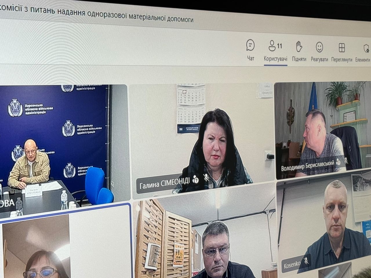 Фотографія екрана комп'ютера, на якому відображено онлайн-нараду у форматі відеоконференції. У верхній частині екрана видно заголовок: «комісії з питань надання одноразової матеріальної допомоги», а також інтерфейс програми з іконками чату та кількістю учасників (11). На екрані розділено кілька вікон із трансляціями учасників: у лівому верхньому кутку видно чоловіка на тлі банера Херсонської ОВА; по центру — жінка з підписом «Галина СІМЕОНІДІ»; праворуч — чоловік у профіль з підписом «Володимир Бериславський». У нижньому рядку помітно вікна інших учасників, зокрема чоловіка в окулярах та чоловіка з підписом «Kosenko».