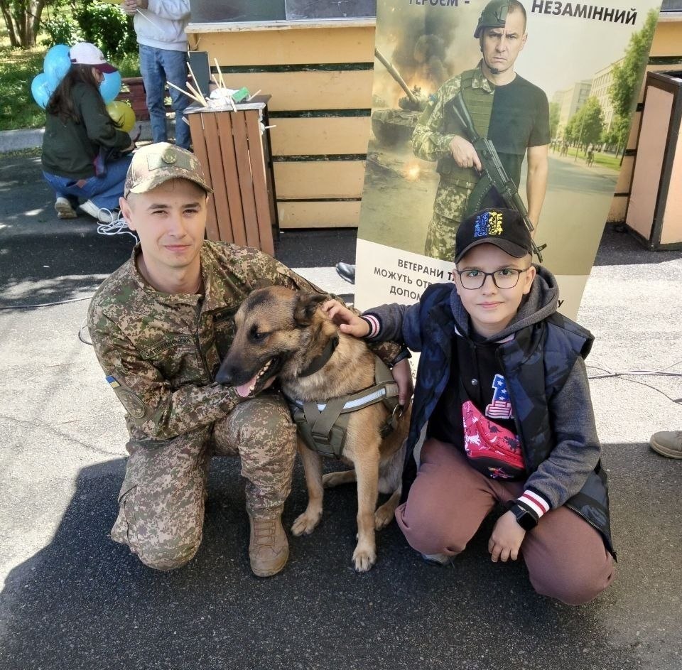 На асфальтованому майданчику військовий у камуфляжній формі та кепці гладить службового собаку в тактичній розгрузці. Поруч із ними присів хлопчик в окулярах та чорній кепці. Позаду стоїть великий банер із зображенням військового на тлі танка та написом про підтримку ветеранів.
