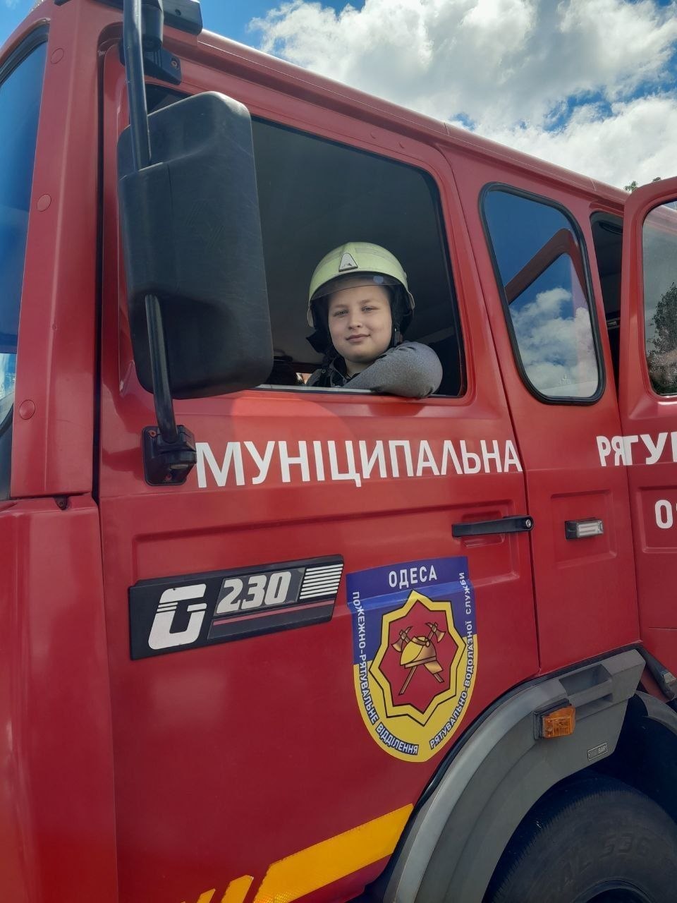 Великий план кабіни червоного пожежного автомобіля. З відчиненого вікна виглядає усміхнений хлопчик у світлому захисному шоломі. На дверях машини розміщено напис «МУНИЦИПАЛЬНА» та емблема пожежно-рятувального відділення міста Одеса.